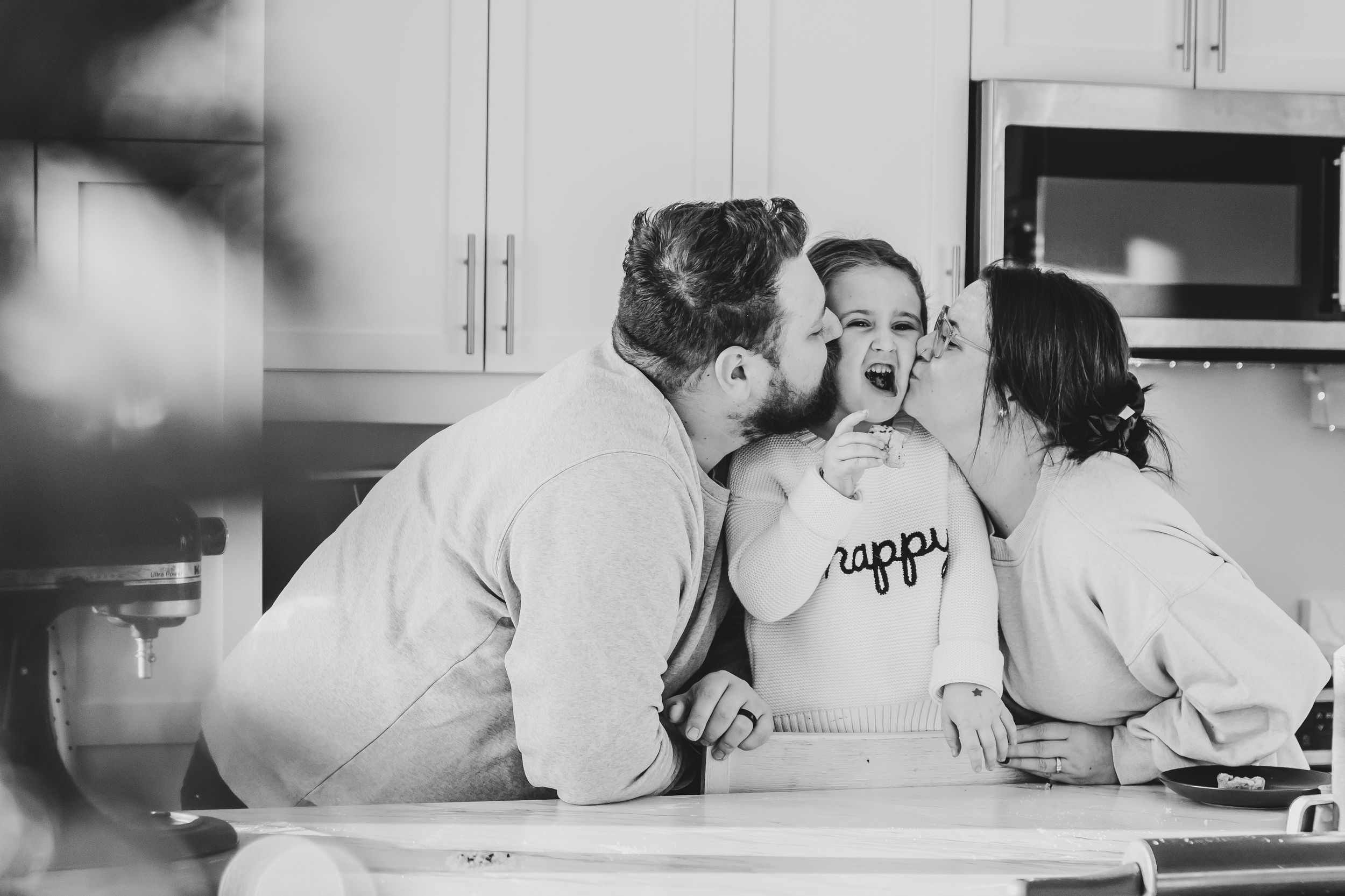 Black and white photo of couple kissing woman on cheeks in bright kitchen.