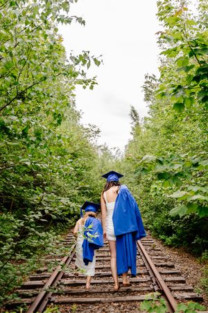 Two graduates in blue caps and gowns walk hand-in-hand down a tree-lined railway track. Scoudouc.