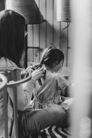 Black and white photo of a woman braiding a young girls hair indoors near a window.