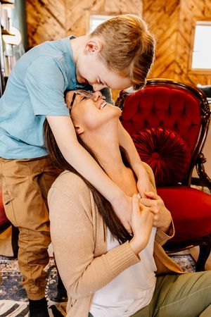 A mother laughs joyfully as her young son leans down to kiss her forehead in a warmly decorated living room.