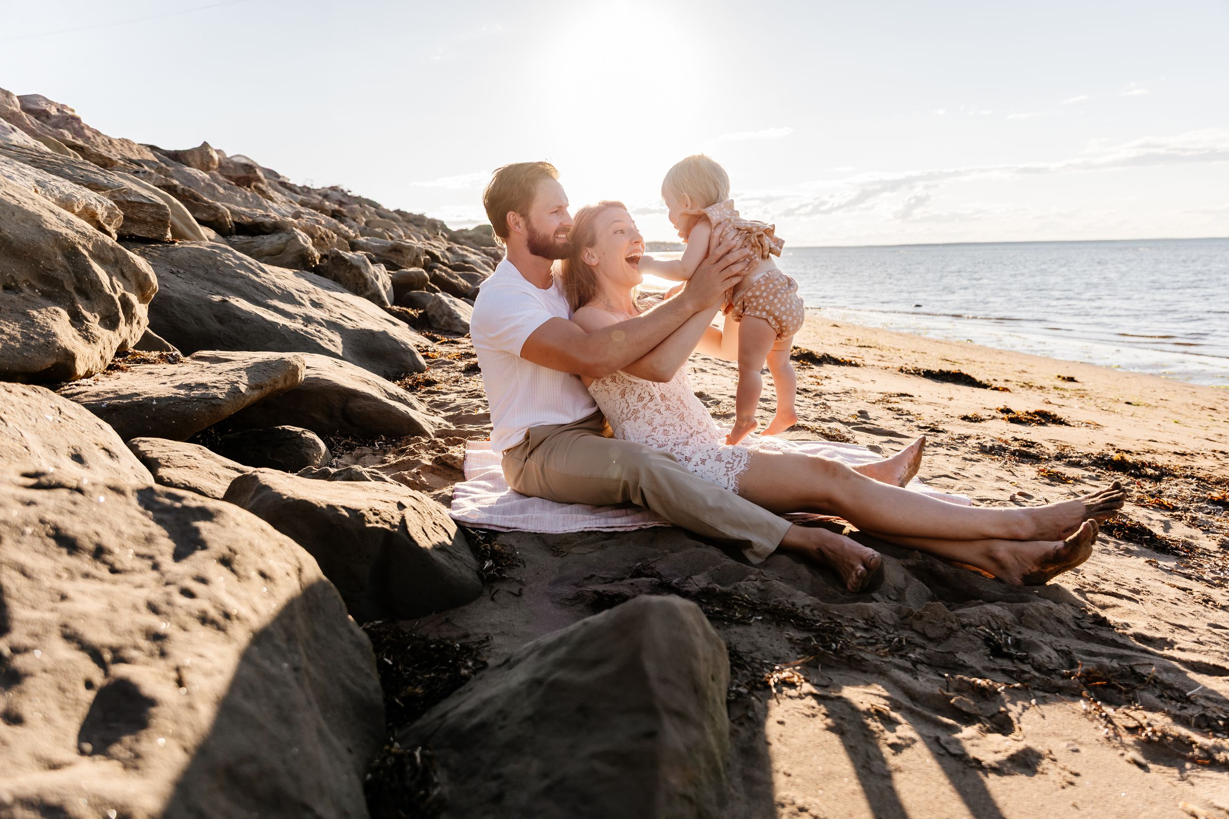 Young couple sitting on rocky beach holding their baby in the air with ocean and sunlight behind them.