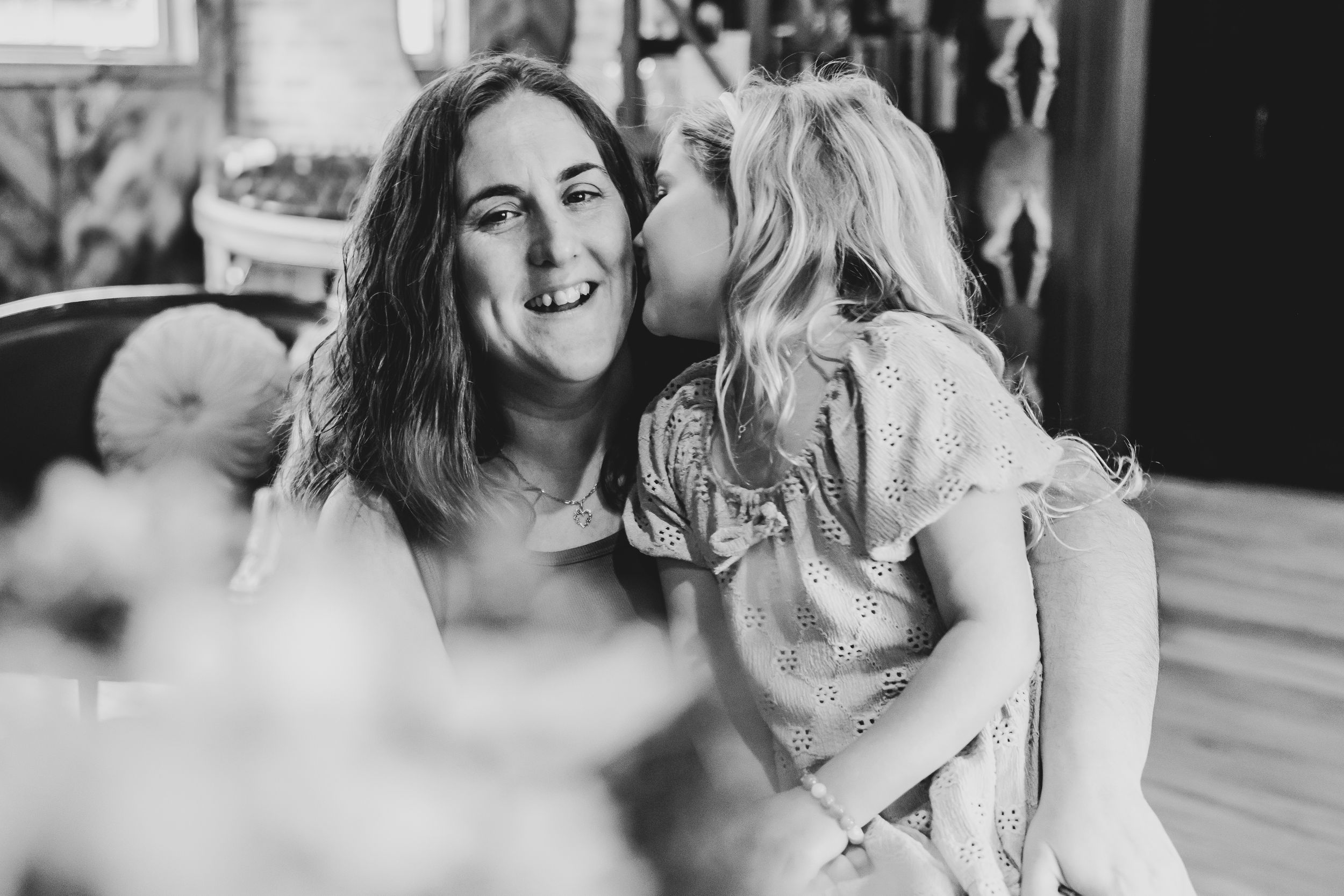 Black and white photo of a young girl kissing her smiling mother on the cheek indoors.