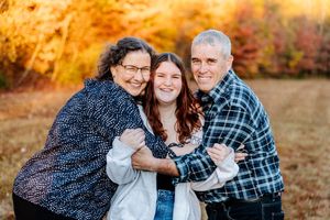 Family of three smiling and embracing outdoors during autumn with colorful fall foliage in the background.