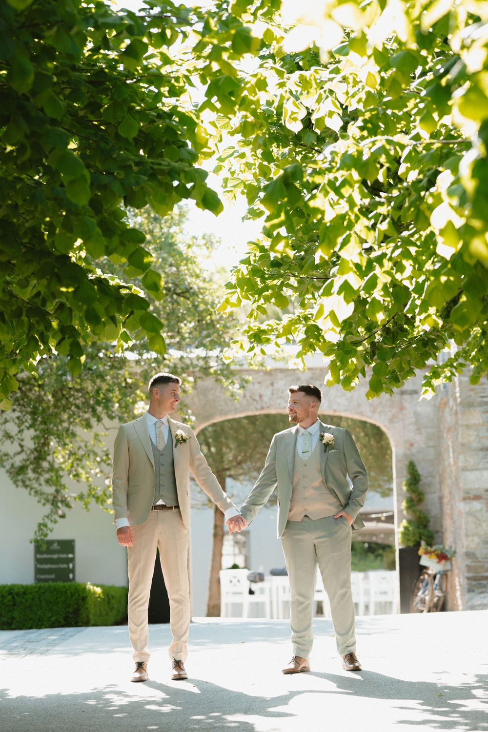 Two grooms in matching sage suits hold hands outdoors, surrounded by lush green trees on their wedding day in Tulfarris Hotel Co.Wicklow