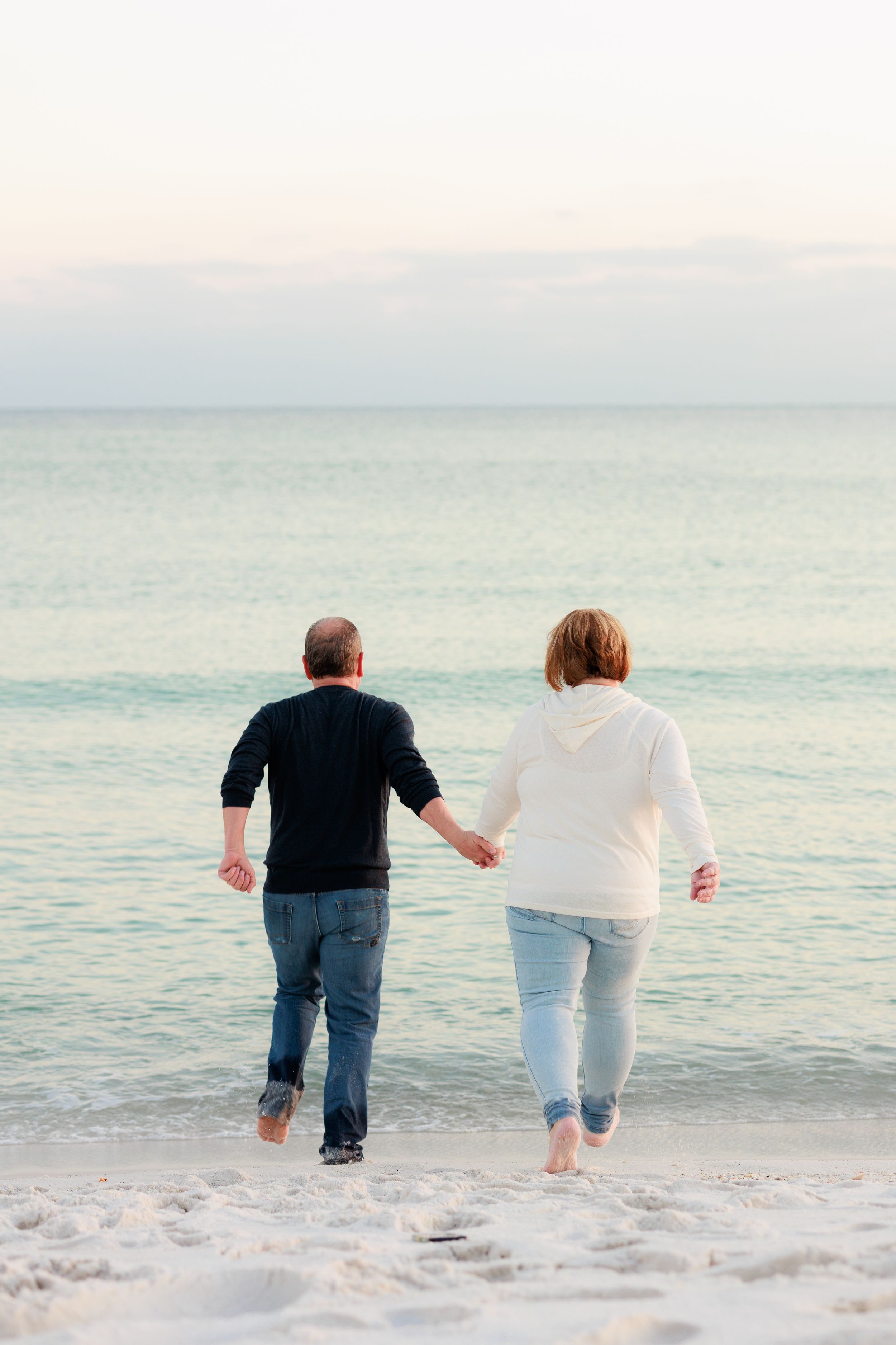 A couple holding hands while walking together on a sandy beach at dusk with calm ocean waters in the background.