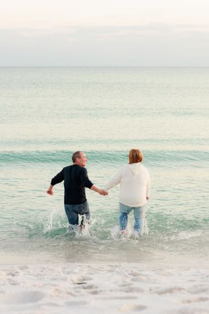 Two people holding hands and splashing in the shallow ocean waves at sunset on a peaceful beach.