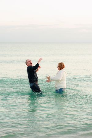 Two people in formal attire playfully fall backwards into turquoise ocean waters during a beach photoshoot.