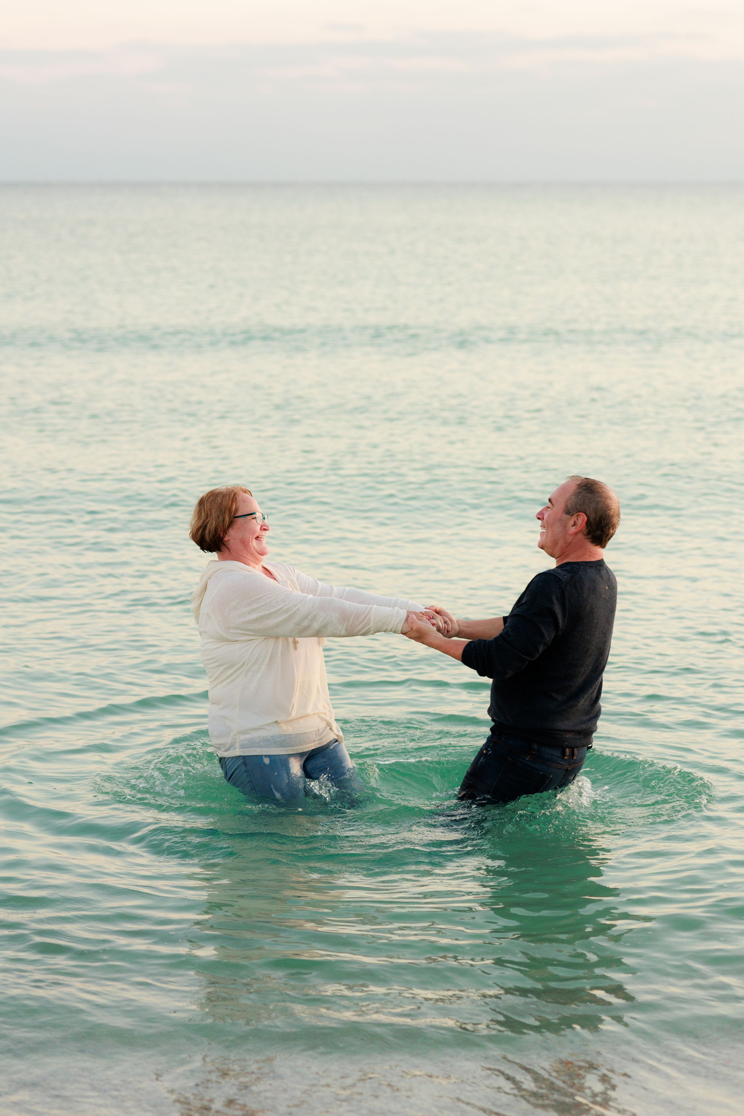 Two people playfully splash and interact in calm turquoise ocean waters during sunset.