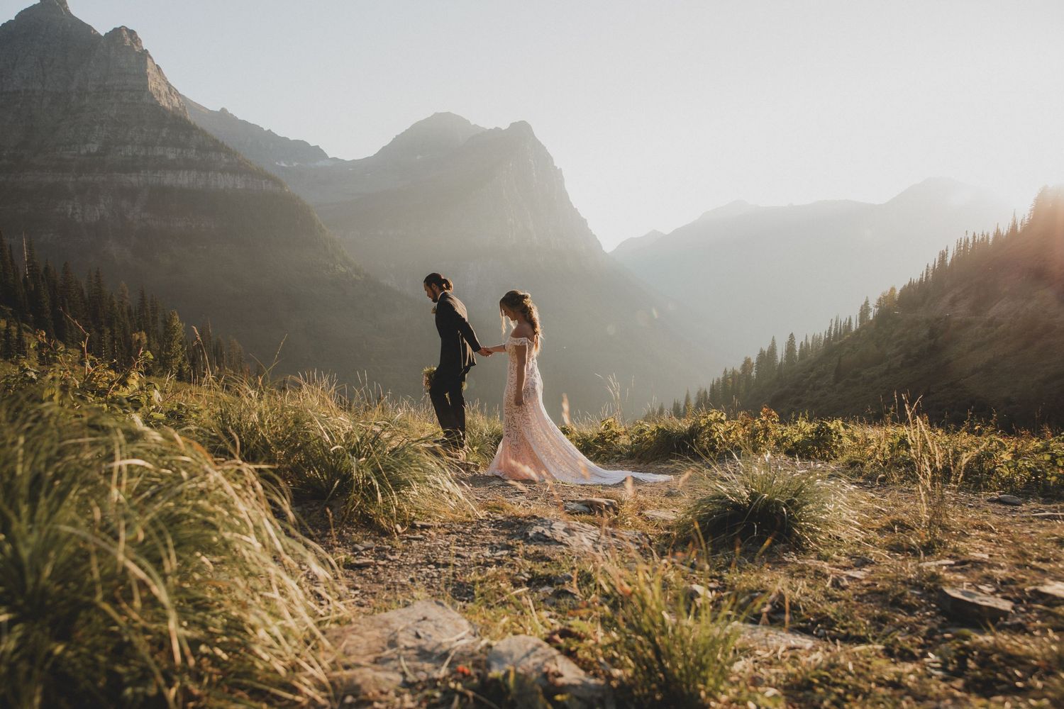 Two figures stand together on a mountain ridge at sunset with dramatic peaks and golden light in Glacier National Park.