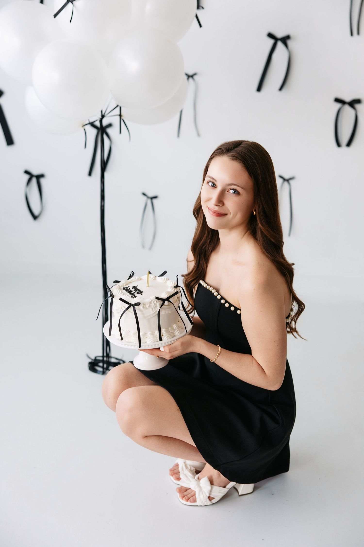 A person in a black dress crouches while holding a gift against a white backdrop with hanging black ribbons.