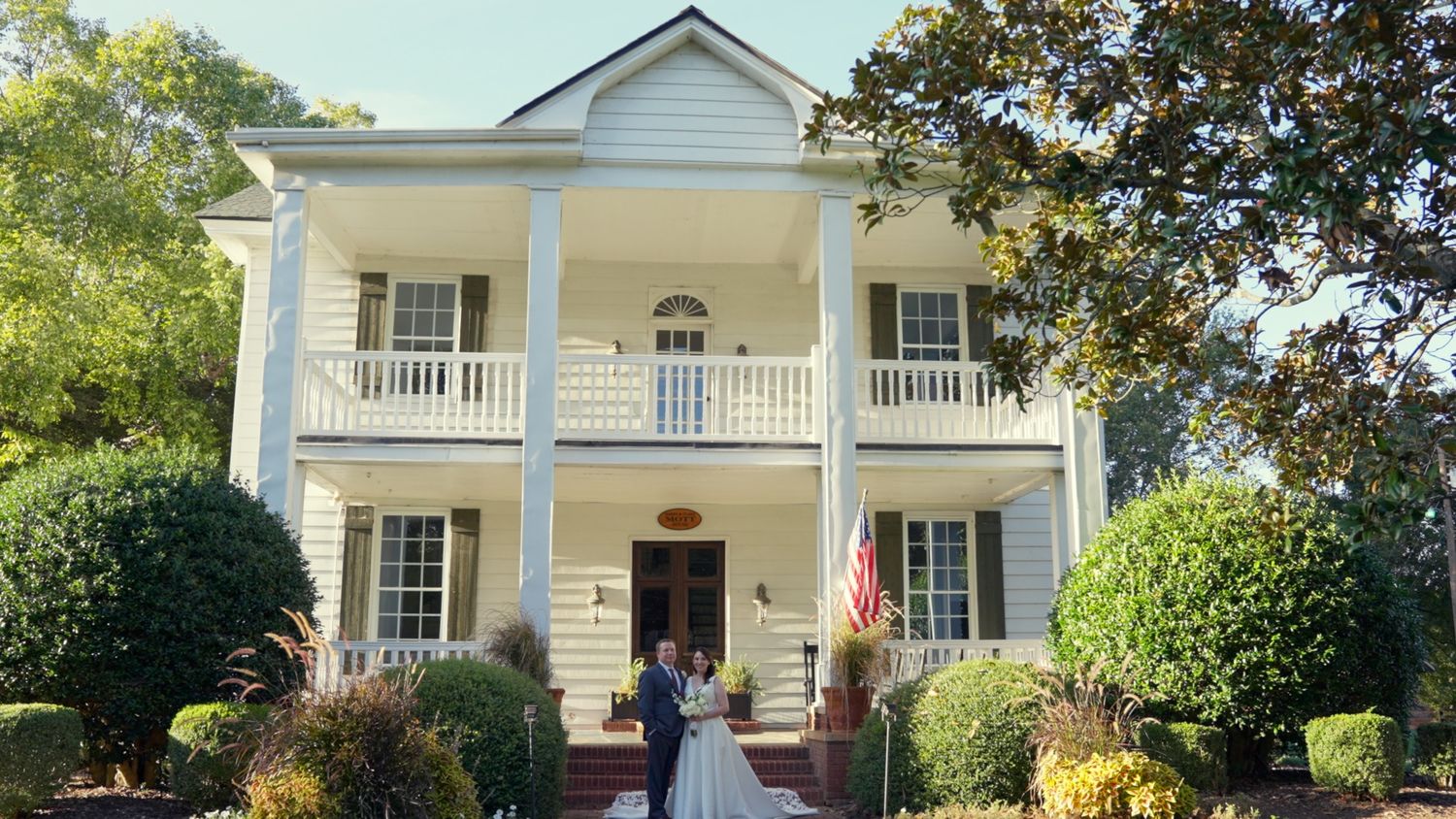 Classic white colonial style house with columns and balcony surrounded by lush greenery on a sunny day.