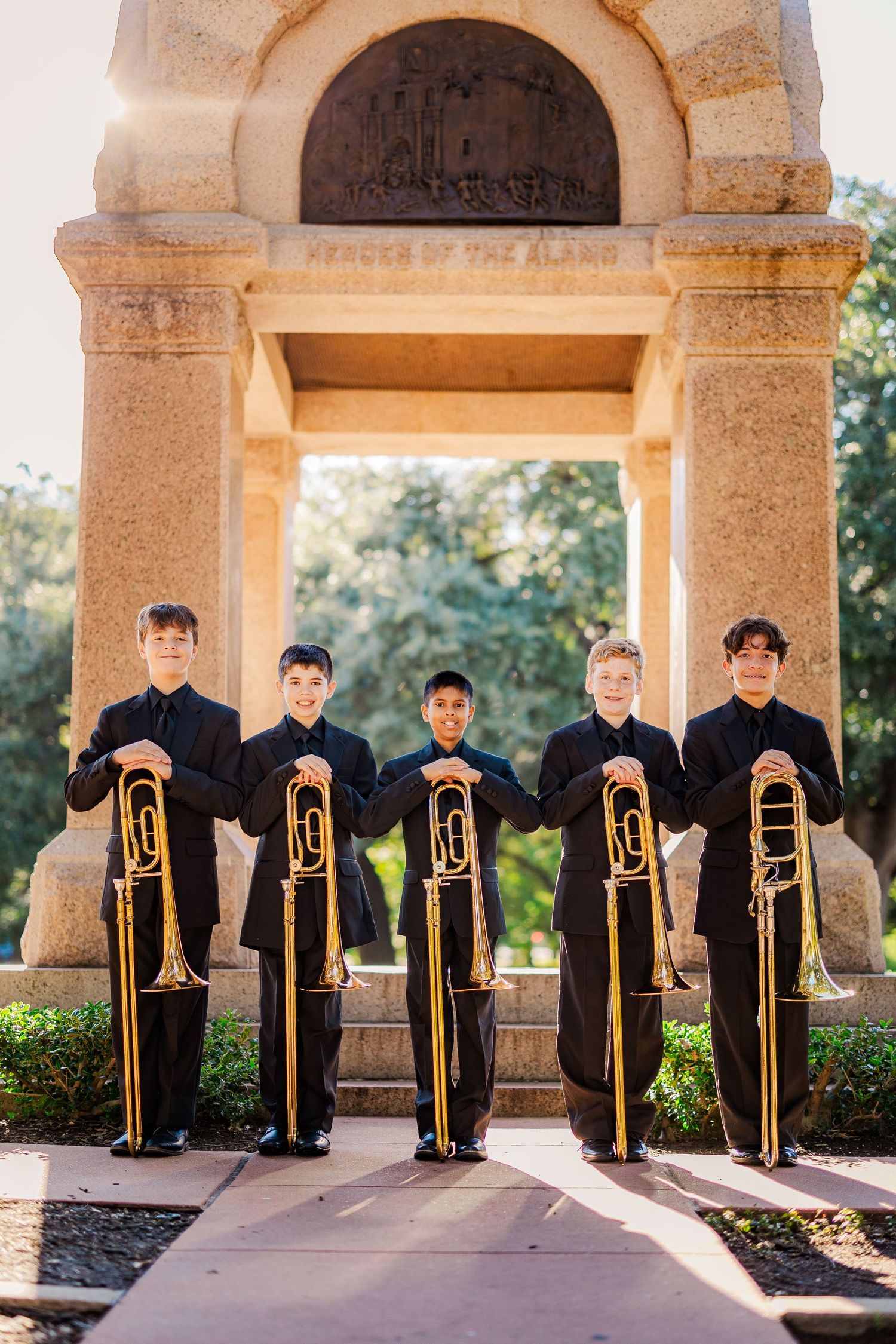 Four young musicians in black uniforms hold trombones before a stone monument in a park setting.