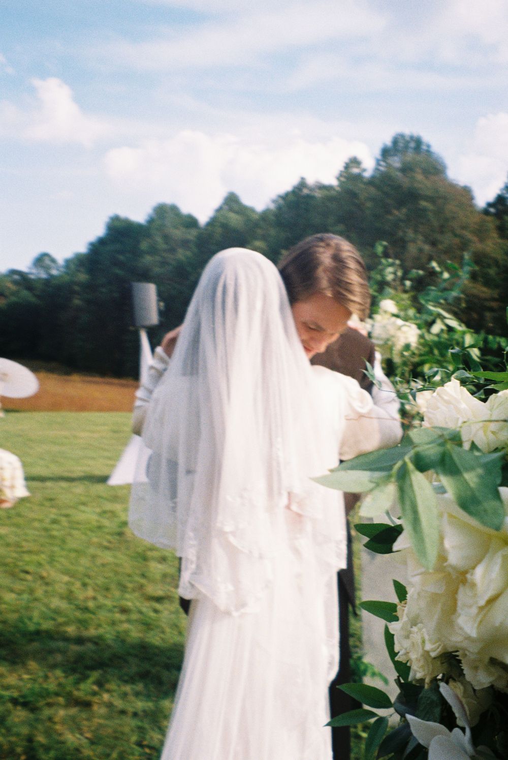 A bride in a flowing white wedding dress and veil stands in a grassy field with white lilies in the foreground.