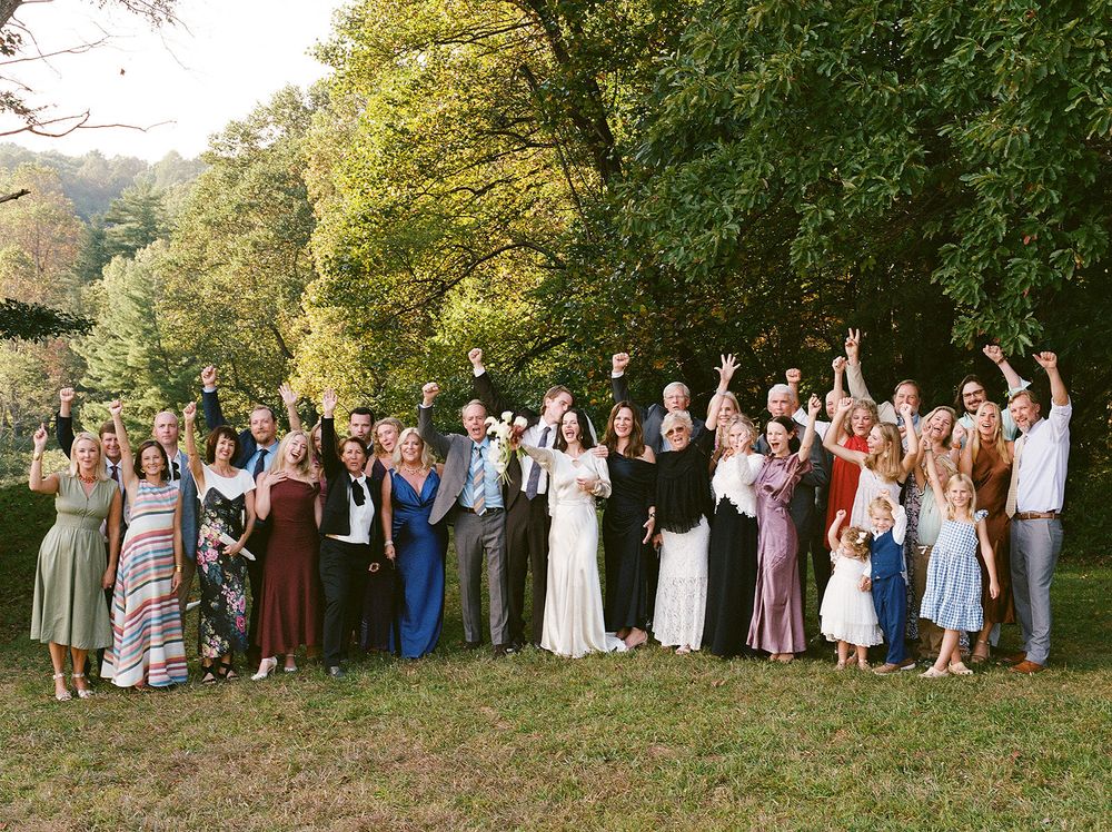 A panoramic group photo of wedding guests lined up outdoors under large trees.