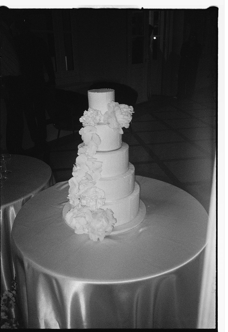 White tiered wedding cake with ruffled floral decorations displayed on a silver tablecloth.