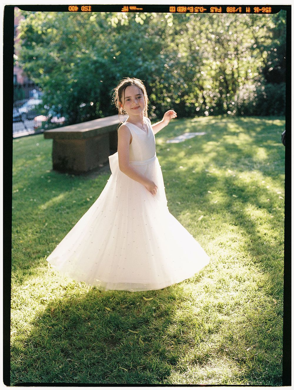 Young flower girl in white dress twirling in sunlit garden setting.