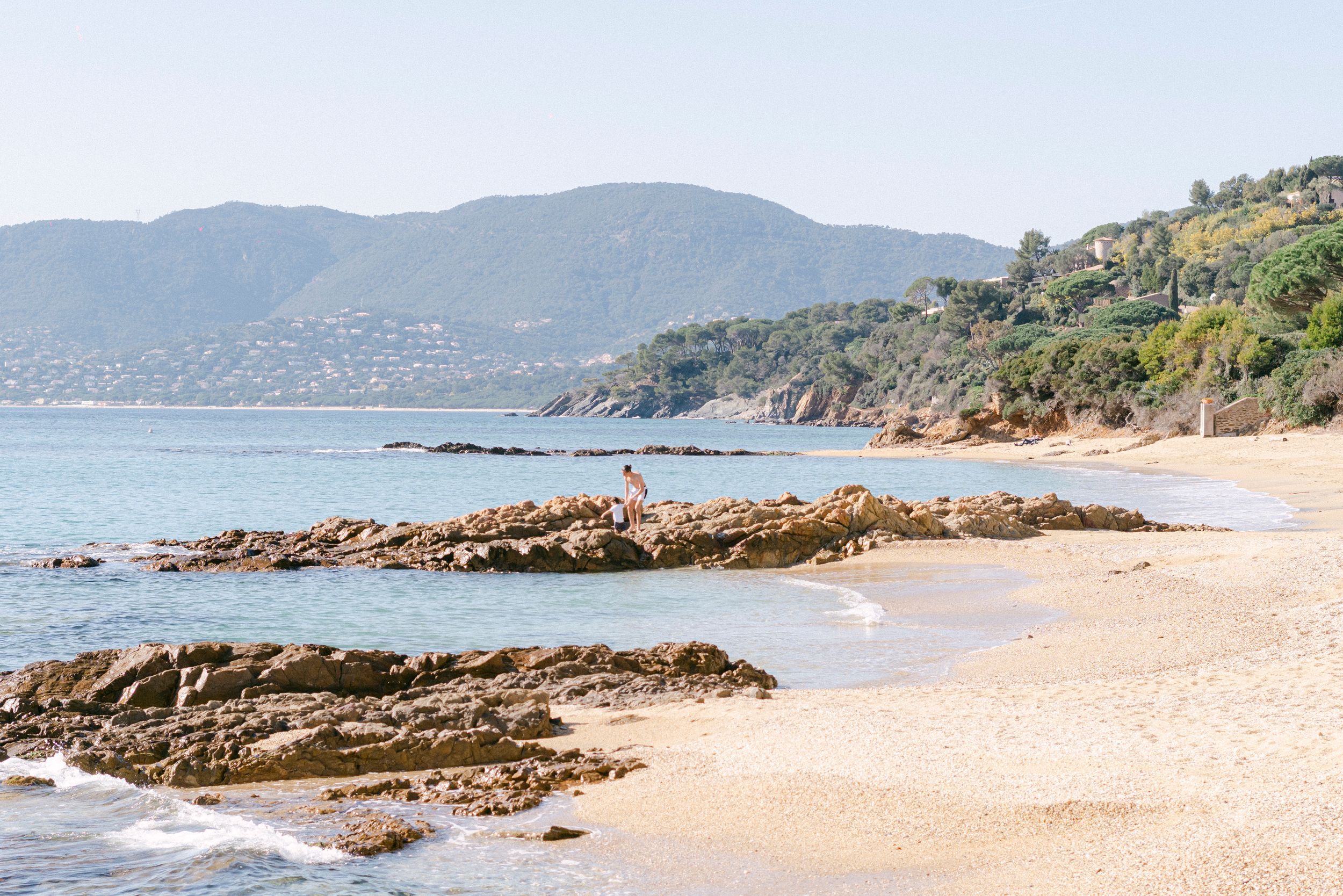 Rocky coastal outcrop stretches along a pristine sandy beach with mountains visible in the background.