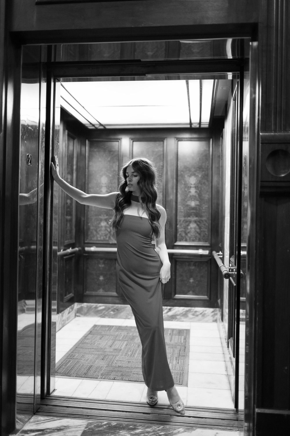 A girl leans up against the door of an elevator dressed very elegantly