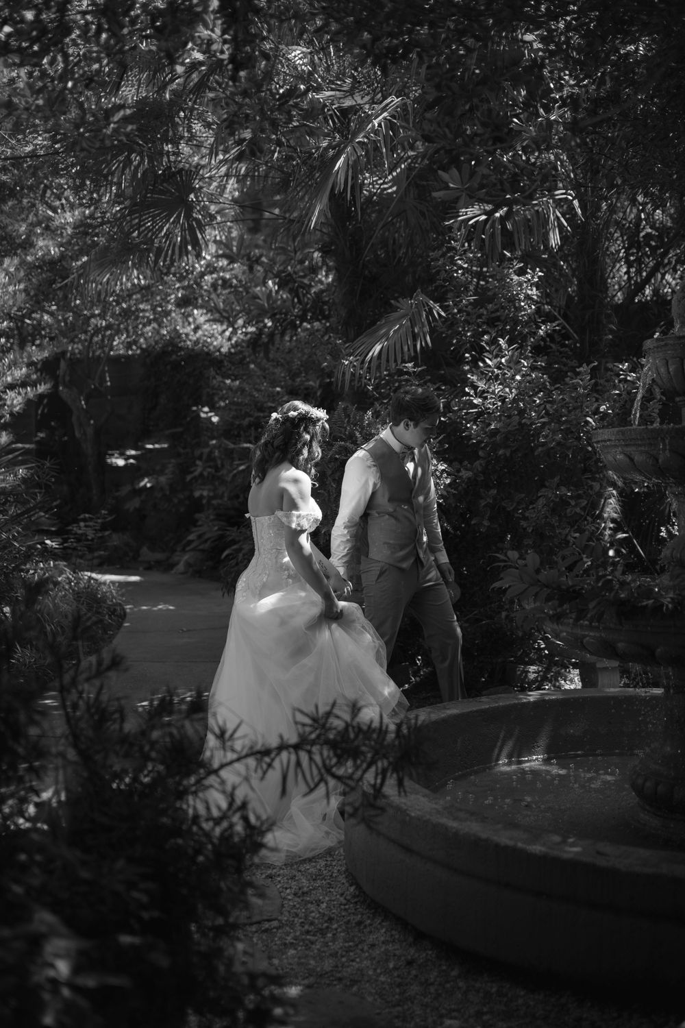 a bride and groom walk holding hands around a fountain