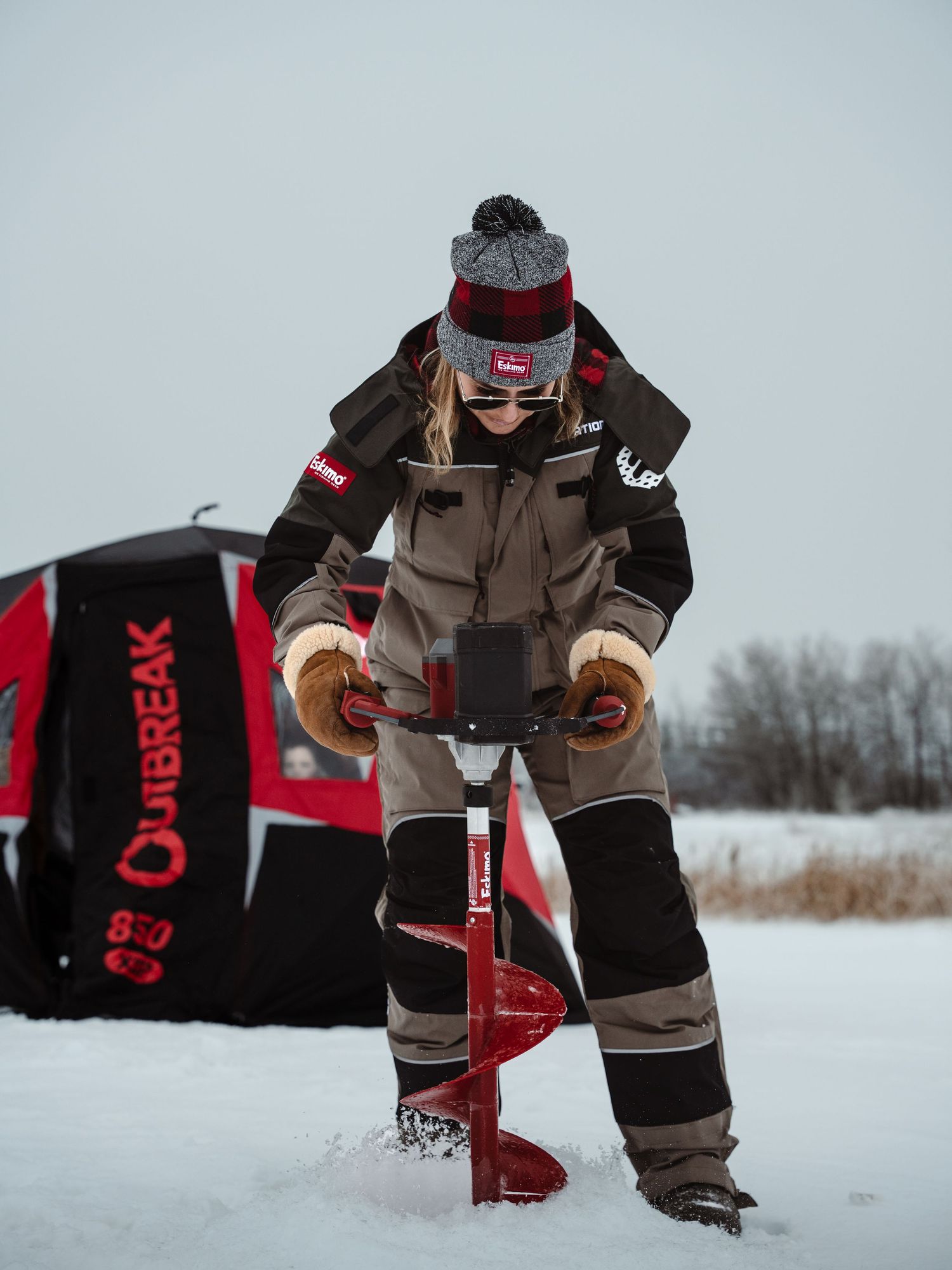 Person in brown winter suit kneeling on snow-covered ice during ice fishing expedition.