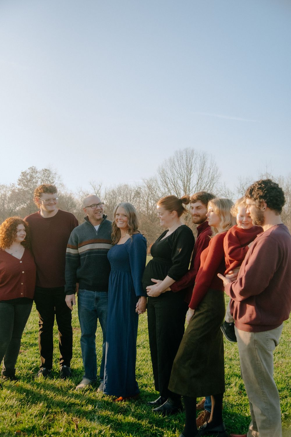 Large group of people standing together in a grassy field during autumn with trees in the background.