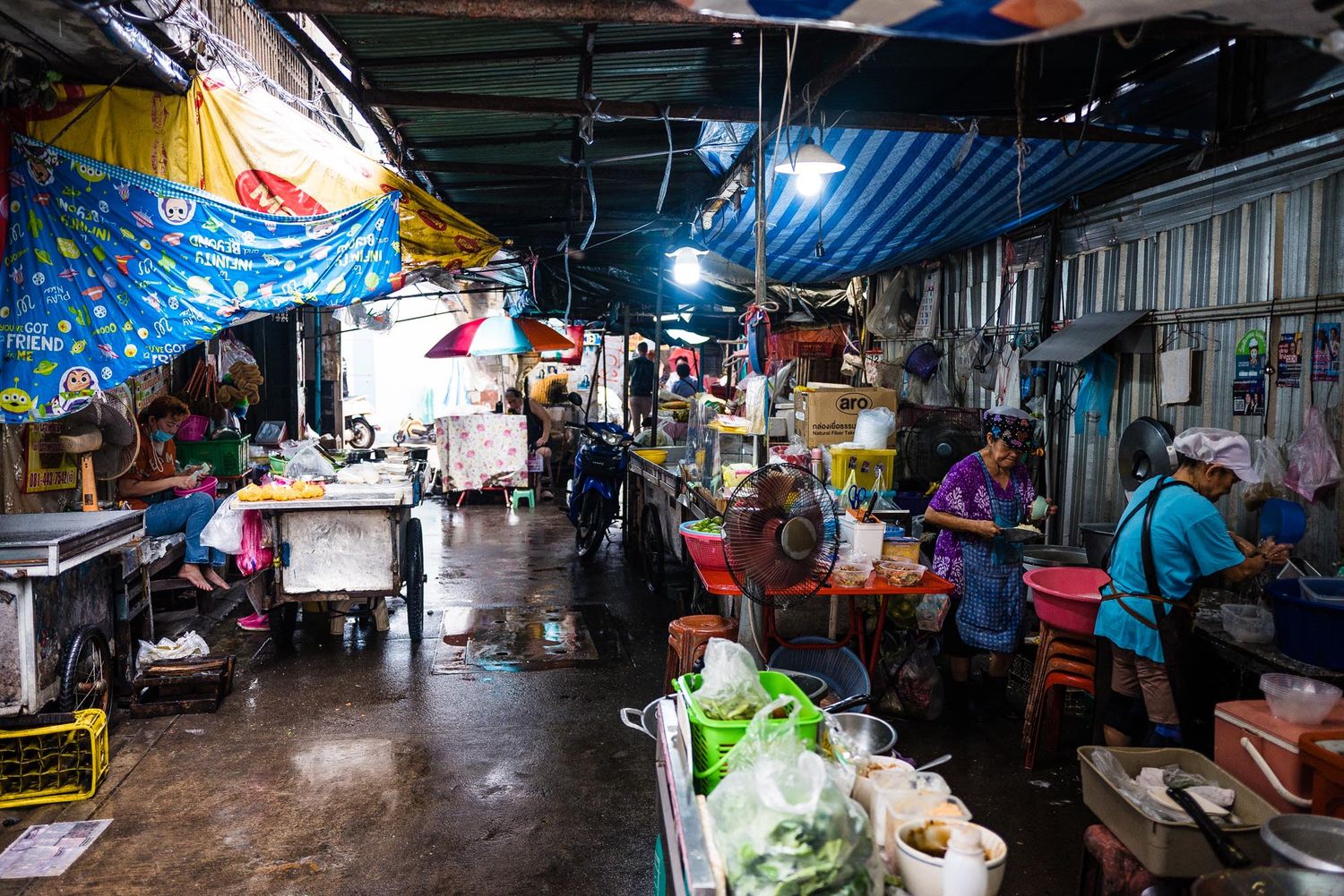 Wet market vendors sell fresh produce and goods in a dimly lit covered marketplace during a rainy day.