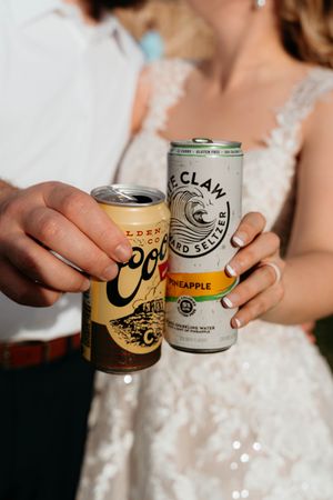 Hands holding various canned beverages in white lace wedding attire against light background.