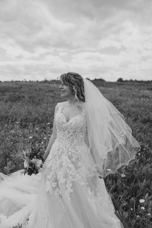 A bride in a lace wedding dress with flowing veil stands in a field of wildflowers on an overcast day.