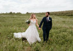 A couple in wedding attire walks hand in hand through a green meadow on their special day.