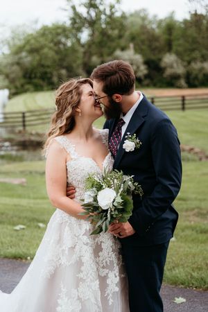 Couple in wedding attire sharing an intimate moment outdoors with white floral bouquet in a pastoral setting.