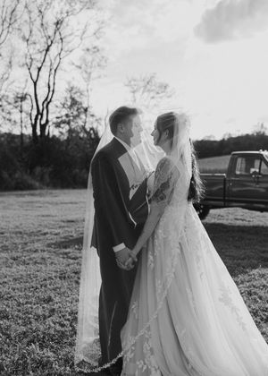 A romantic black and white wedding photo shows a couple in formal attire standing together outdoors on a grassy field.