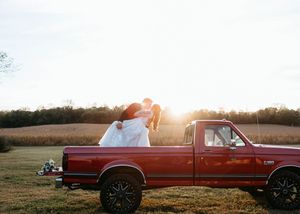 Couple embraces on red pickup truck in countryside field during golden sunset.
