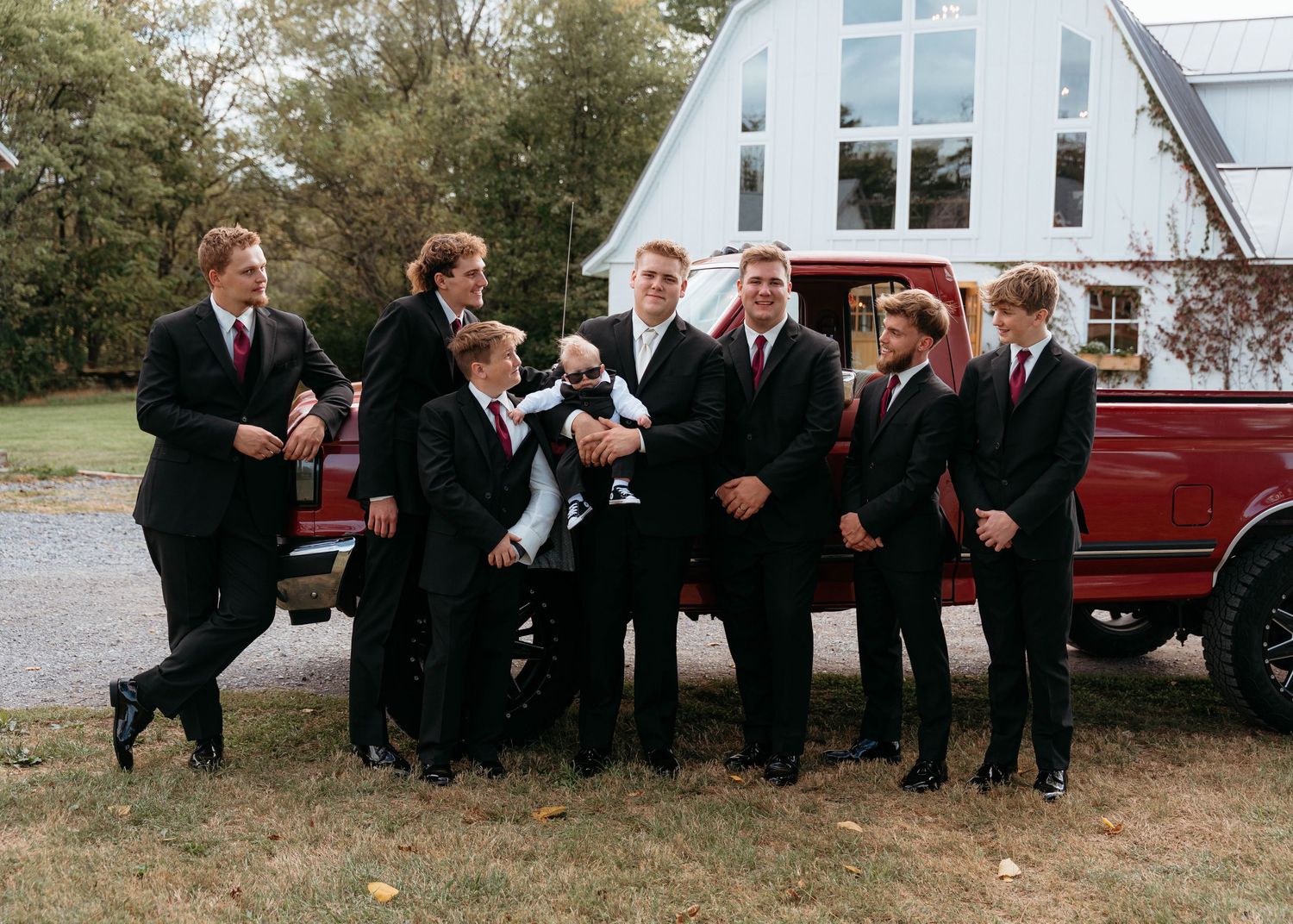 A group of groomsmen in black suits pose playfully near a red vintage truck outside a white A-frame building.