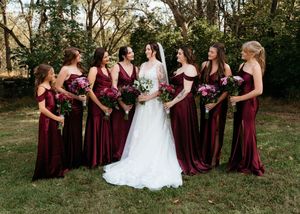 A group in burgundy dresses and a white gown pose together for outdoor wedding photos in a natural setting.