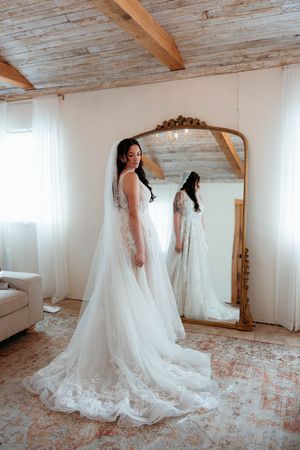 A flowing white wedding gown with a long train and veil displayed in a rustic room with wooden beams and floral carpet.