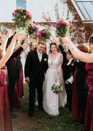 Wedding party in burgundy attire forms celebratory arch with raised glasses as couple walks beneath on their special day.