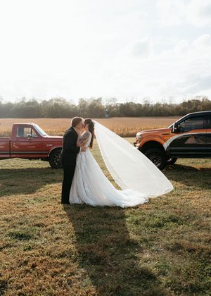 A wedding couple poses in a rustic field at sunset with vintage cars in the background.