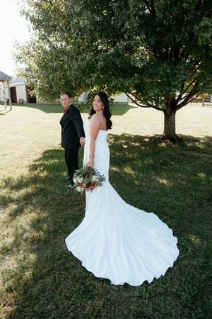 A romantic wedding couple poses outdoors under trees on a grassy lawn, with the bride's white dress flowing elegantly.