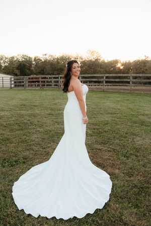 A bride in a flowing white mermaid wedding gown poses in a grassy field at sunset with a rustic fence in background.