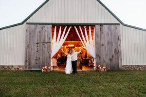 A rustic barn wedding venue with white drapes framing the entrance and warm glowing lights inside at dusk.
