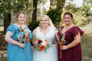 Wedding party members hold vibrant orange and burgundy floral bouquets in a woodland setting.