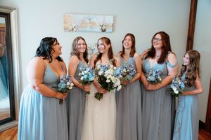 A group of bridesmaids in light blue and gray dresses hold bouquets while standing together at a wedding.