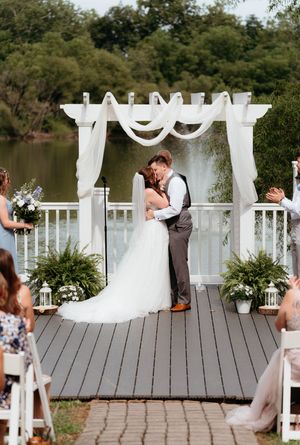 A romantic wedding couple shares a tender moment on a lakeside deck with large decorative white letters in the background.