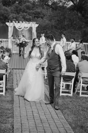 A black and white outdoor wedding ceremony with a couple walking down a brick path past white chairs and floral arch.