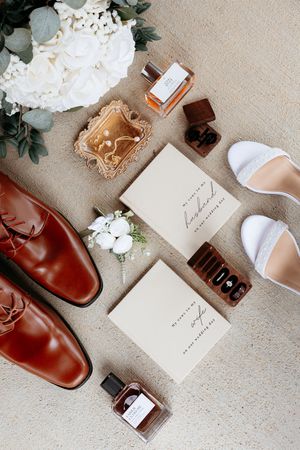 Elegant wedding flat lay with brown leather shoes, white heels, perfume bottles, boutonniere, jewelry, and vow books on neutral background.