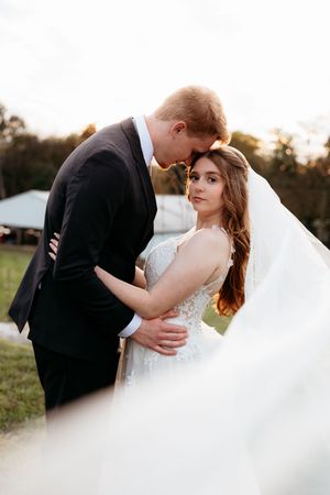 A bride and groom share a romantic embrace outdoors at golden hour, her veil flowing in the breeze.