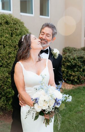 A joyful wedding couple share a candid laughing moment while holding a white and blue floral bouquet outdoors.
