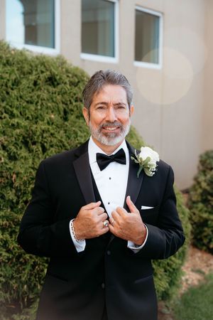 A person in a black tuxedo and bowtie adjusts their collar while smiling outdoors.