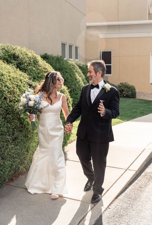 A bride and groom share a playful moment walking together outdoors while holding drinks in formal wedding attire.