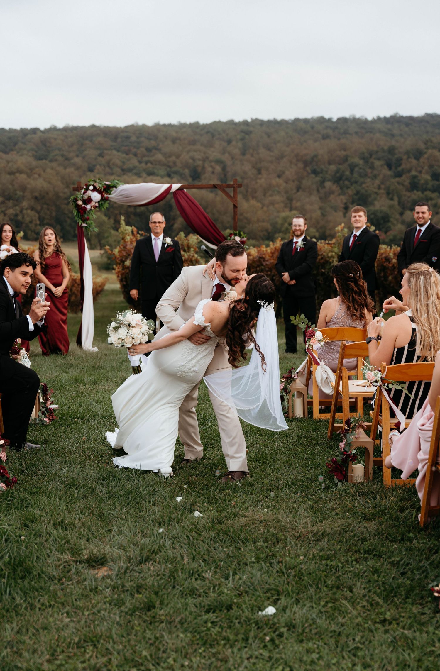 Wedding guests seated on wooden chairs in an outdoor ceremony space with rolling hills in the background during autumn.